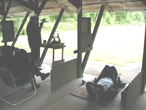 Image of three people: first person is standing while aiming a big bore silhouette. Clockwise next person is laying on his back while shooting big bore silhouette. Next and final person is sitting in a chair watching.