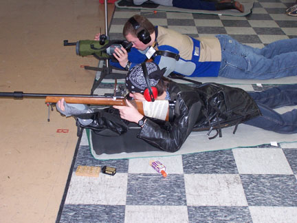 Image of two teenagers laying on the ground. The teenager in the foreground of the image is aiming a wooden small bore rifle and the teenager in the background is looking through a green scope.