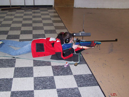 Image of a teenager wearing a red jacket laying on the floor while aiming a blue small bore rifle.