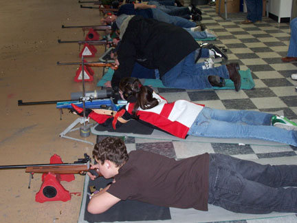 Image of teenagers laying on the floor in a row while aiming a variety of small bore rifles. A teenager in the middle of the row is receiving guidance from an instructor on aiming.