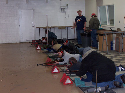 Image of teenagers laying on the floor in a row while aiming a variety of small bore rifles. Two adult instructors are standing in the back corner while supervising.