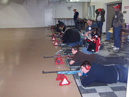 Image of teenagers laying on the floor in a row while shooting a variety of small bore rifles. One teenager wearing a red jacket is receiving guidance from an instructor. Three other instructors are supervising behind the row.