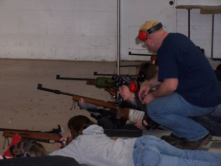 Image of an instructor crouching on the floor while guiding a teenager. The teenager is laying on the floor in a row with other teenagers aiming a small bore rifle.